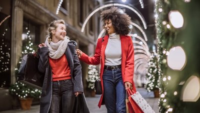 صديقات شابات جميلات يبتسمن على نطاق واسع بطريقة مرحة ويلهون في مركز تسوق، ويشترين هدايا عيد الميلاد